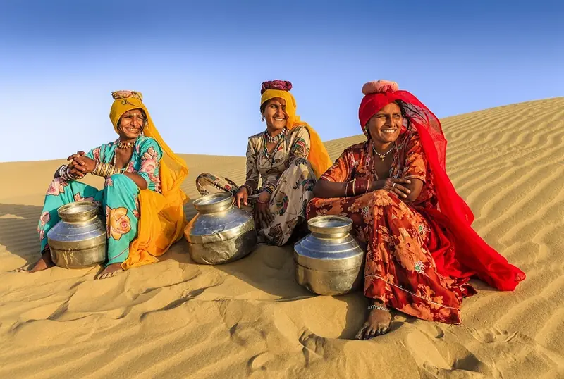 Rajasthani women in bright clothes sitting on desert dunes with brass pots
